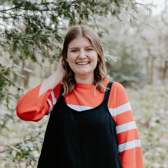 Zabby Allen is a nature enthusiast and designer from south east London. The photo shows Zabby, a white woman with dark blonde hair in her early 30s, stands under a tree. She is wearing an orange and white striped jumper and black dungarees.